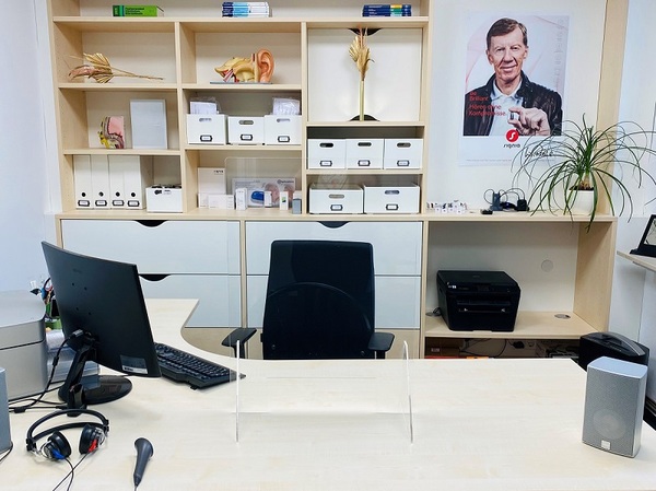 Modernes Hörakustiker-Büro mit Schreibtisch, Computer, Regalen und Hörgeräte-Zubehör.